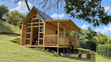 Una cabaña de madera con balcón, ubicada en una ladera cubierta de hierba y árboles al fondo.