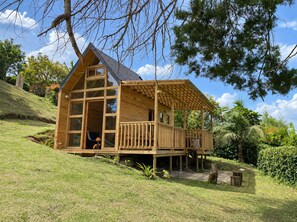 A wooden cabin with a balcony, situated on a grassy slope with trees in the background.