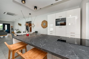 A modern kitchen with a marble countertop, wooden bar stools, and stainless steel appliances. - Casa Romelli • Luxury 5BR Family Villa • Pool, Whirlpool & Tropical Garden by Bocobay (Noord)