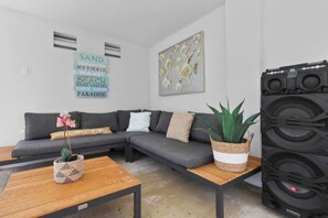A modern living room with a sectional sofa, a coffee table, a potted plant, a wall-mounted speaker, and decorative wall art. - Casa Romelli • Luxury 5BR Family Villa • Pool, Whirlpool & Tropical Garden by Bocobay (Noord)
