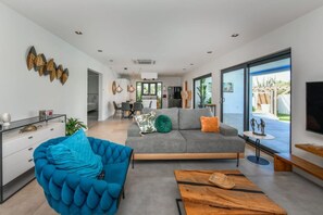Ein modernes Wohnzimmer mit einer Couch, einem blauen Sessel und einem Couchtisch. Im Hintergrund ist ein Essbereich mit Tisch und Stühlen zu sehen.