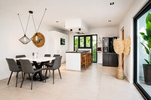 Een moderne keuken met een centraal eiland, een eetgedeelte met een witte tafel en stoelen, en een groot raam met uitzicht op groen.