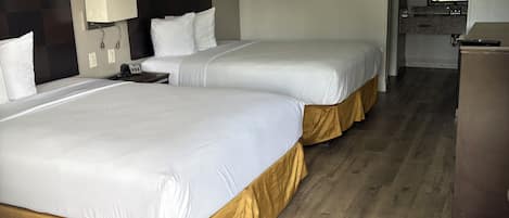 Quarto de hotel com duas camas, uma escrivaninha, luminária e uma cozinha compacta.