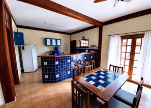 A kitchen with a blue tiled backsplash, wooden cabinets, a refrigerator, and a dining area with a table and chairs. - House with 4 suites, 100m from the beach | Pool & Gourmet Kitchen | 1.2km from the village (Ilhabela)