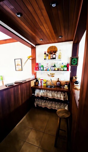 A bar area with a wooden ceiling, a bar counter, and a stool. There are bottles and glasses on the counter, and a clock on the wall. - House with 4 suites, 100m from the beach | Pool & Gourmet Kitchen | 1.2km from the village (Ilhabela)