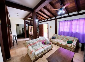 A living room with a wooden ceiling, a sofa, a coffee table, and a dining area with chairs and a table. - House with 4 suites, 100m from the beach | Pool & Gourmet Kitchen | 1.2km from the village (Ilhabela)