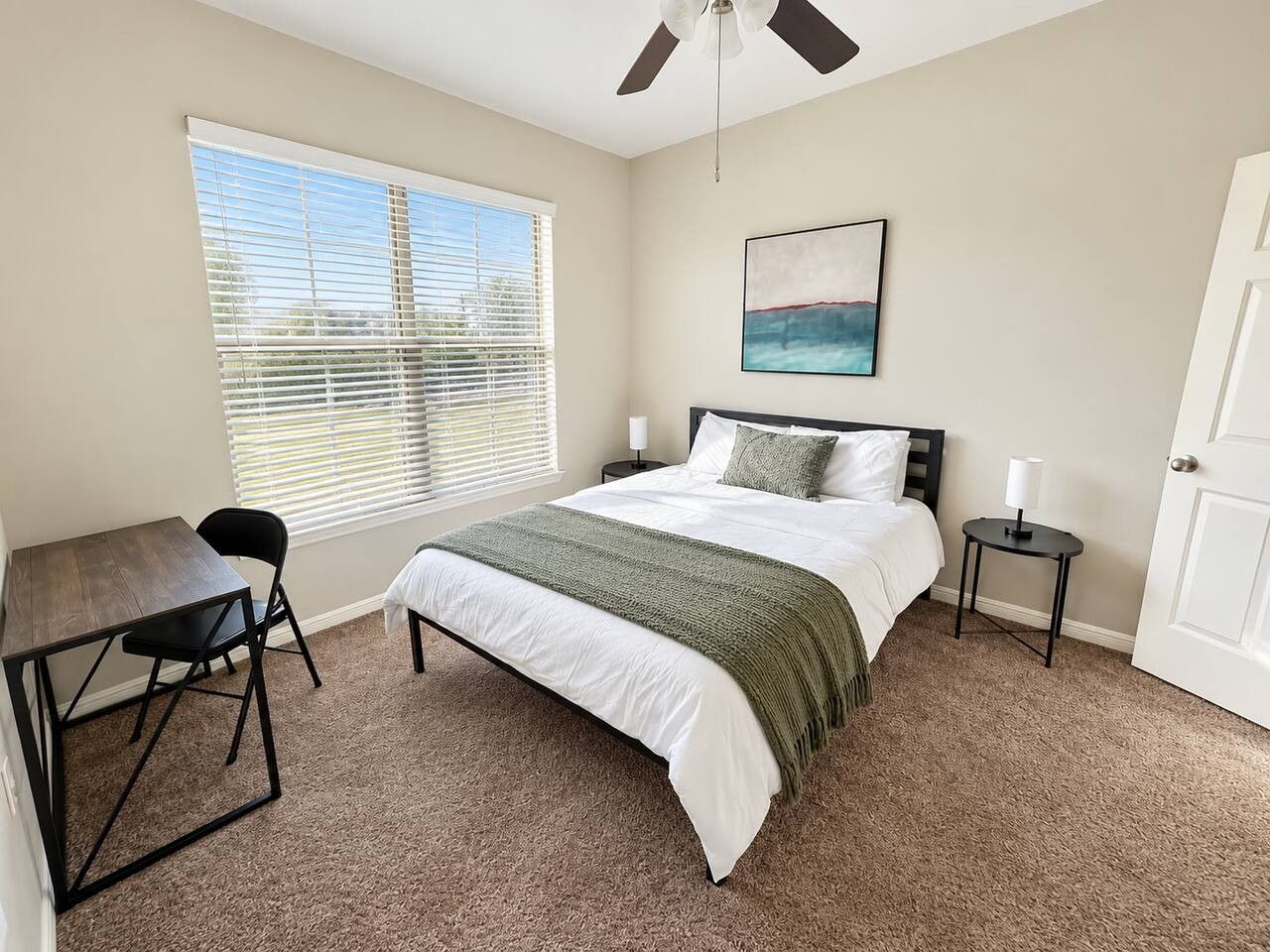 A bedroom with a bed, a desk, a chair, a nightstand, a lamp, a ceiling fan, and a window with blinds.