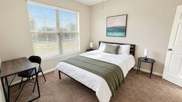 A bedroom with a bed, a desk, a chair, a nightstand, a lamp, a ceiling fan, and a window with blinds.