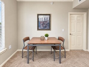 A dining area with a wooden table and four chairs, a framed picture on the wall, and a door to the right. - Sleepover | Breezy 1BD/1BA + Gym - Corpus Christi (Corpus Christi)