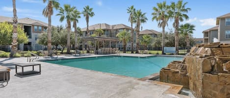 A swimming pool with lounge chairs and palm trees, surrounded by buildings.