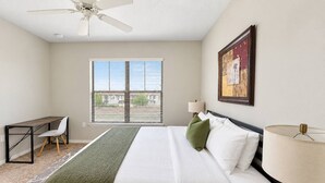 Une chambre d’hôtel comprenant un lit, un bureau, une chaise, une fenêtre avec des stores, un ventilateur de plafond et une lampe fixée au mur.