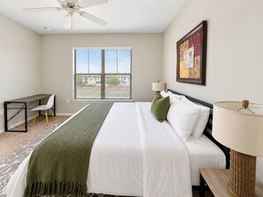 A hotel room with a bed, a desk, a chair, a window with blinds, a ceiling fan, and a wall-mounted lamp. - Sleepover | Breezy 1BD/1BA + Gym - Corpus Christi (Corpus Christi)