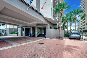 A modern building with a red-brick pathway, a flag, and palm trees. - Beach Colony 210 - Steps to the Beach (Myrtle Beach)