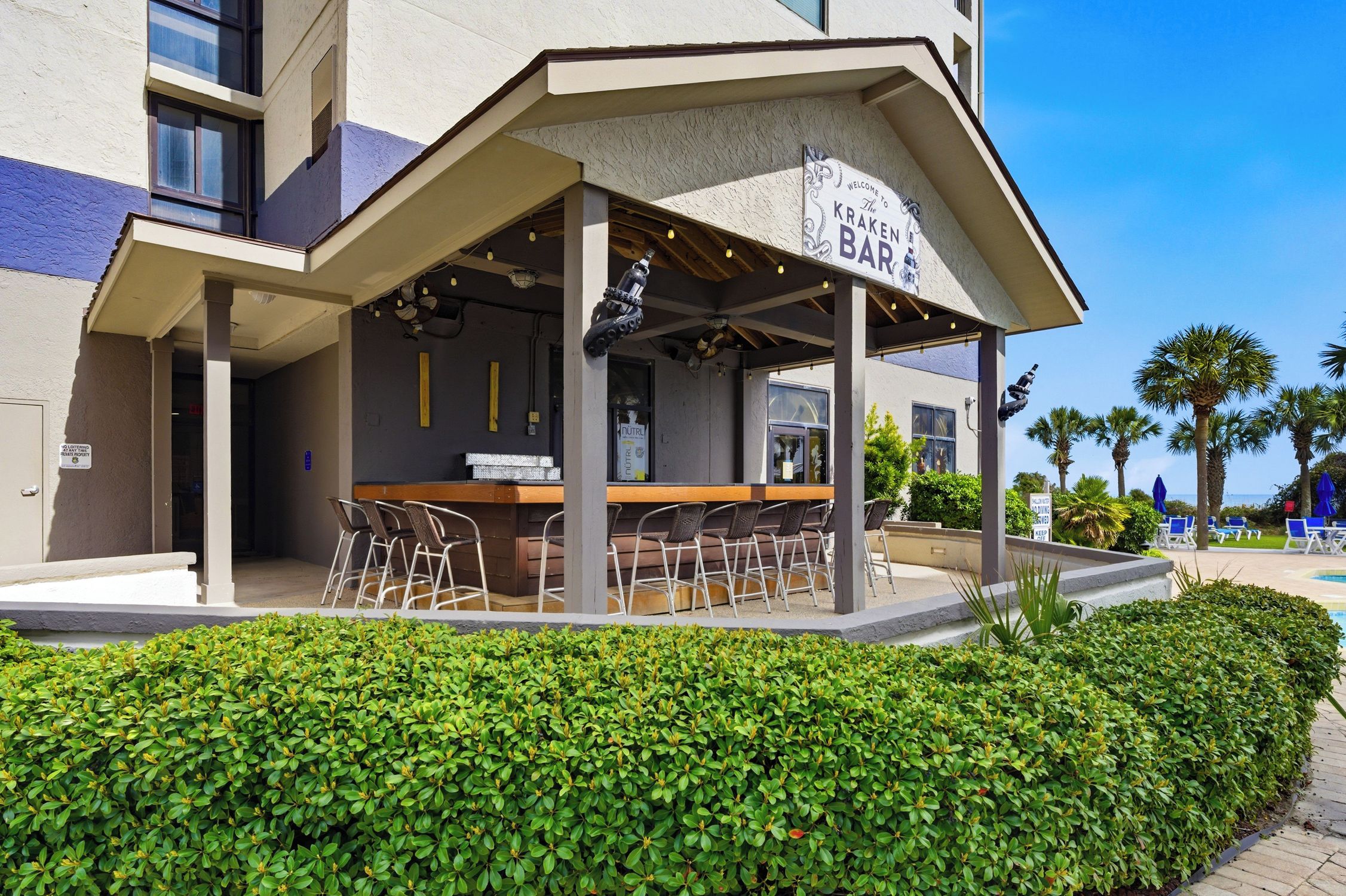 A bar with outdoor seating, a sign reading 'Welcome to the Kraken Bar,' and a pool area with palm trees in the background.