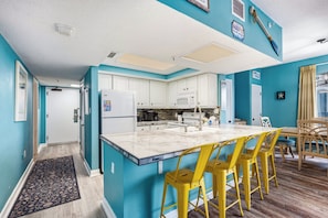 A kitchen with white cabinets, a large island with bar stools, and a view of the living area. - Beach Colony 210 - Steps to the Beach (Myrtle Beach)