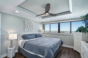 A bedroom with a bed, a ceiling fan, a window with blinds, a decorative wall piece, a bedside lamp, and a dresser. - Holiday Towers 1103 - Oceanview 3BR Condo (Myrtle Beach)