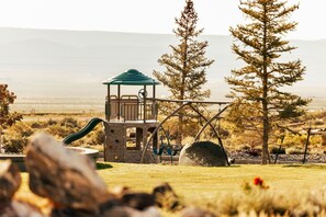 A playground with a slide and climbing area, surrounded by trees and a grassy field.