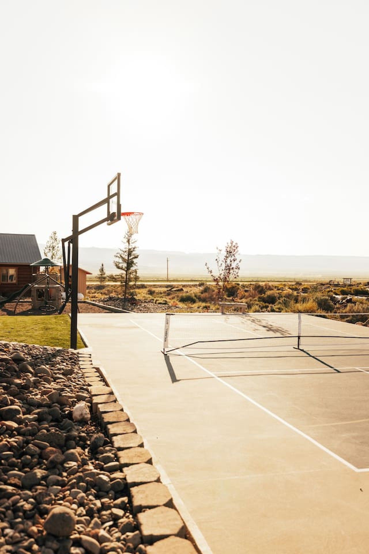 Una cancha de baloncesto al aire libre con un aro, una estructura de juegos de madera y una casa al fondo.