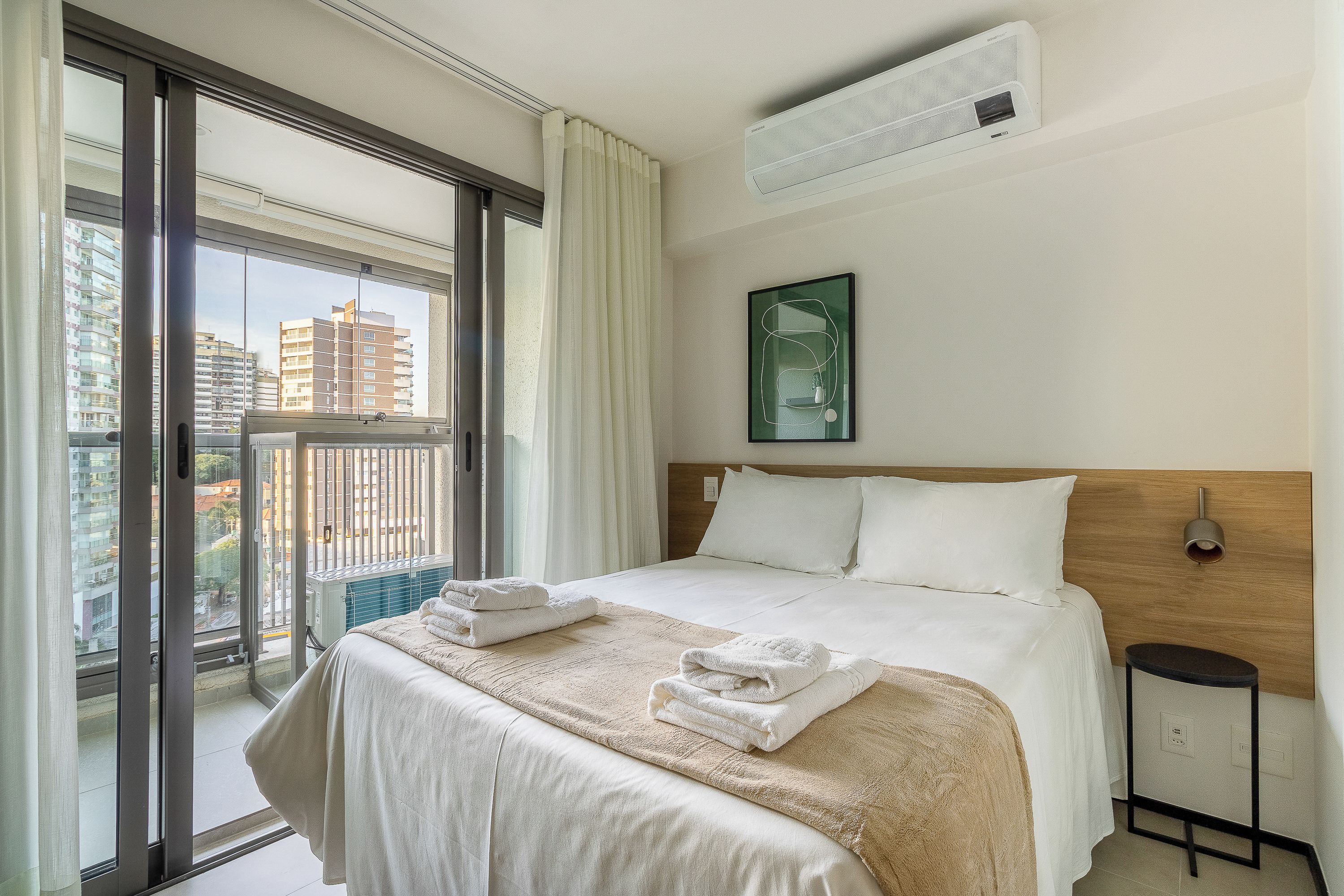 A hotel room with a large bed, a window with a view of buildings, and an air conditioning unit on the wall.