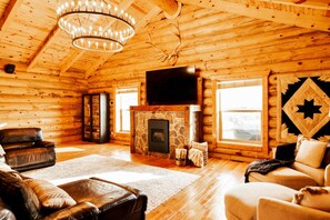 A log cabin interior with a stone fireplace, leather furniture, and a large flat-screen TV mounted above it.