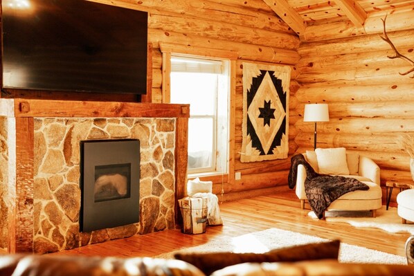 A cozy log cabin interior with a stone fireplace, a mounted television, a patterned rug, and a comfortable seating area.