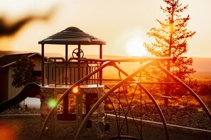 A playground with a swing set and a slide, surrounded by trees and a sunset in the background.
