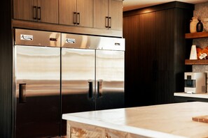 A modern kitchen with stainless steel double-door refrigerators, wooden cabinets, and a marble countertop.