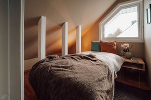 A bedroom with a sloped ceiling, a bed with a brown blanket, a nightstand with a vase and book, and a window with a view. - Kaare’s Villa in Tromsø - Guest Favorite (Tromsø)