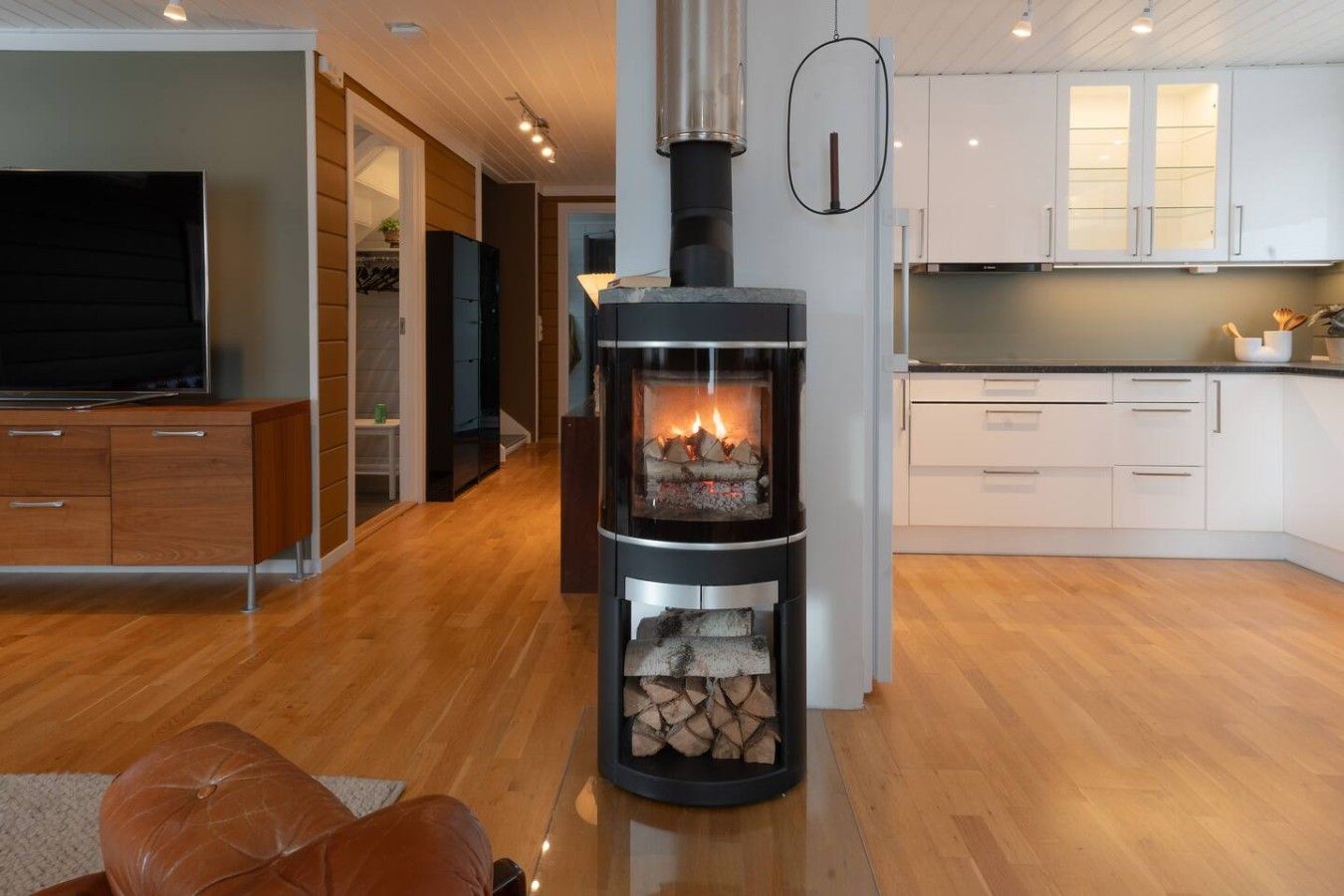 A modern living room with a wood-burning stove, a TV, and a kitchen area with white cabinets.