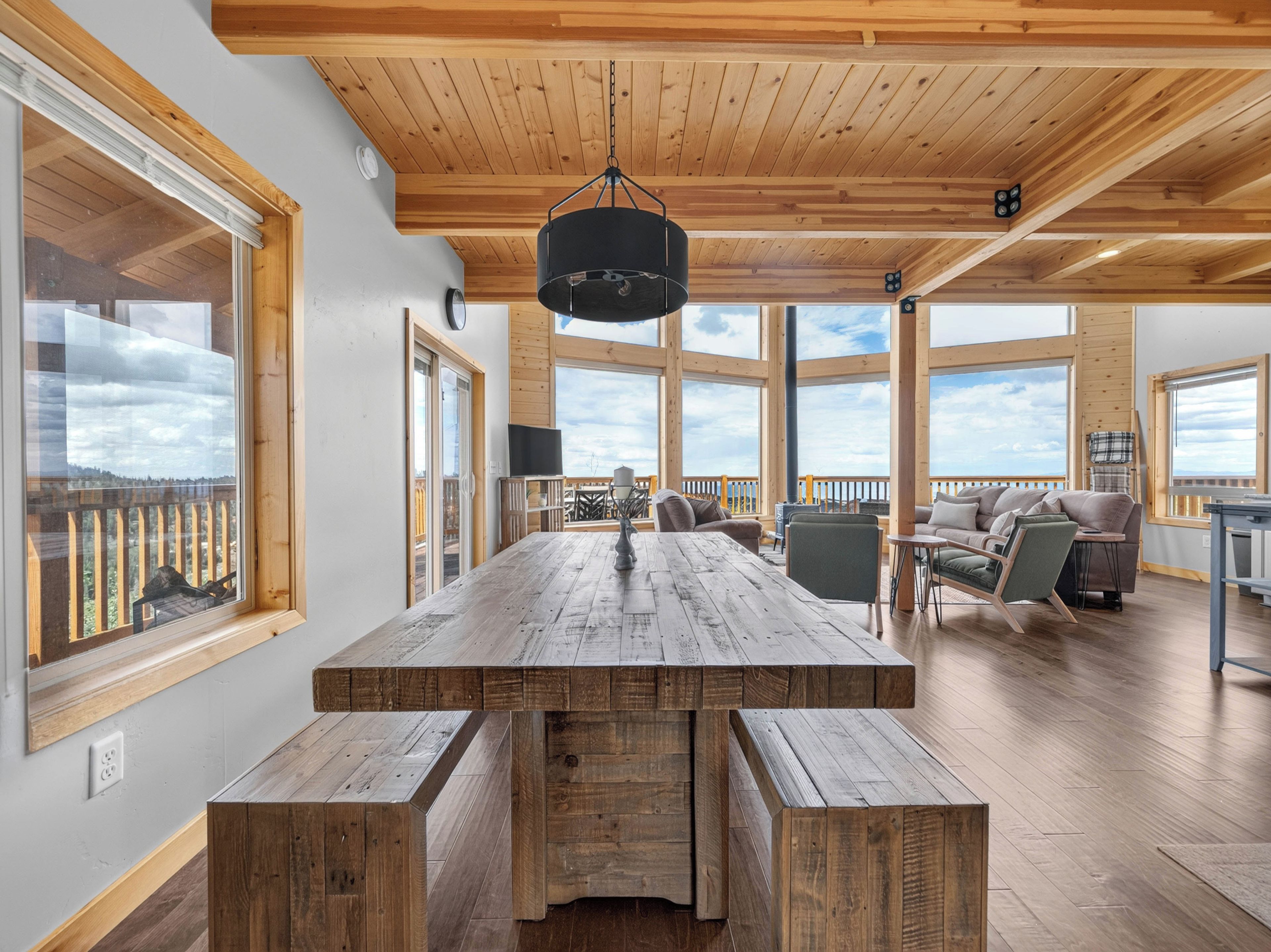 A spacious room with a large wooden table and benches, large windows offering views, and a ceiling with wooden beams.