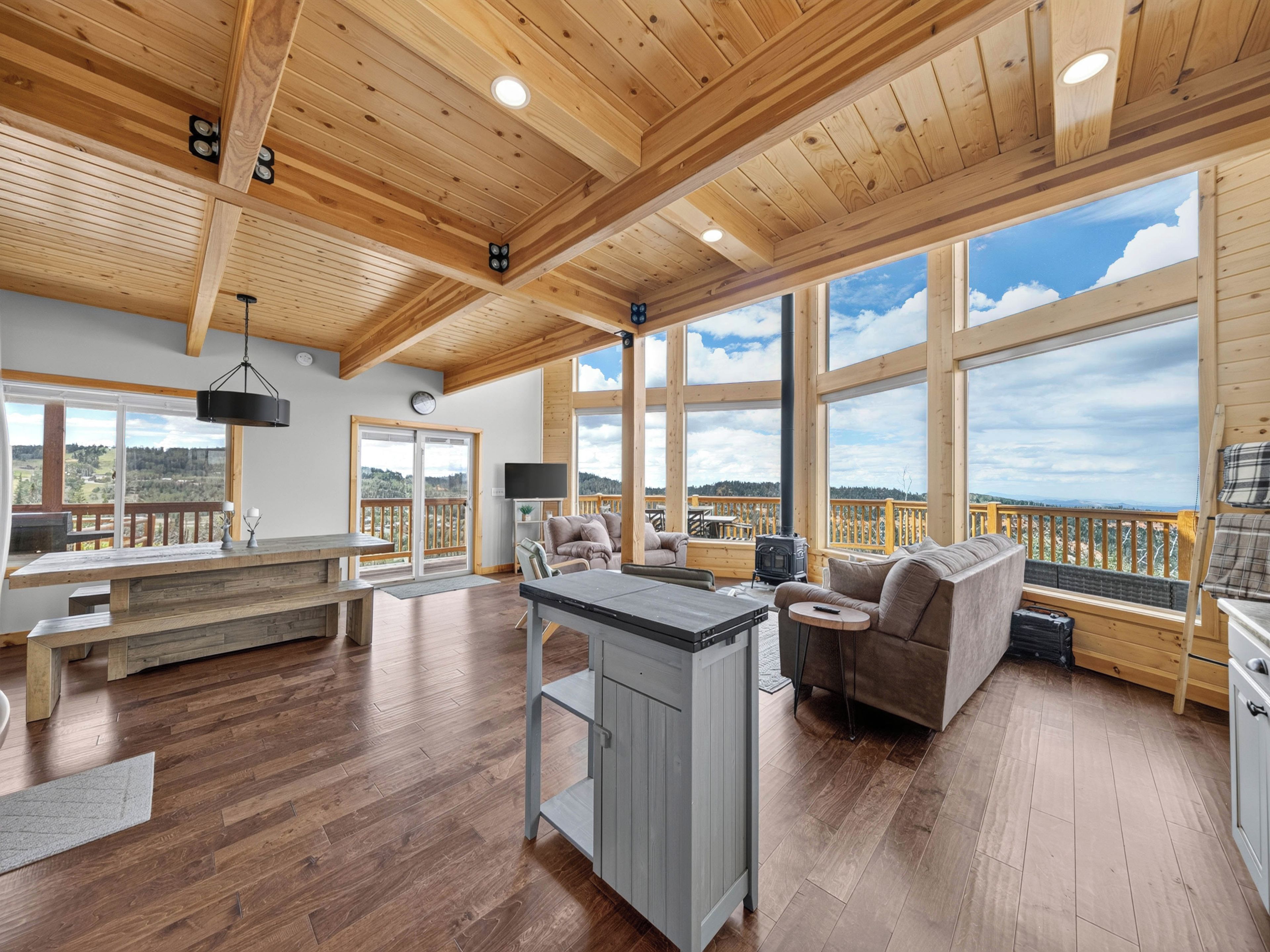 A spacious living area with a wooden ceiling, large windows, a fireplace, and a kitchen area.