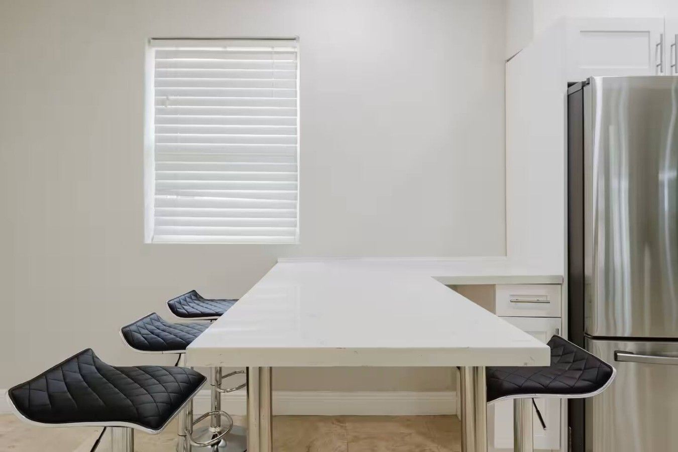 A kitchen with a white countertop, a stainless steel refrigerator, and a window with blinds.