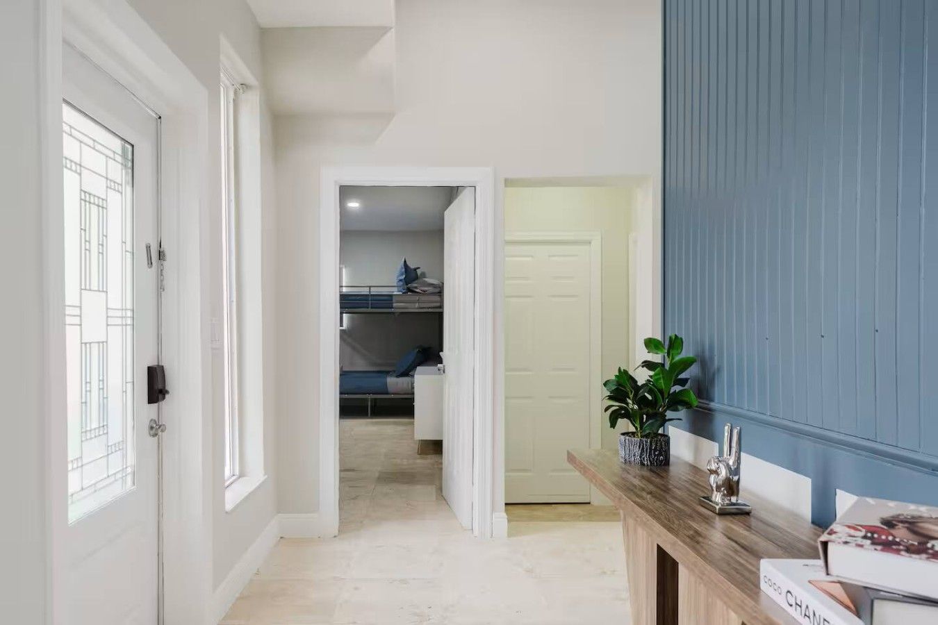 Un couloir avec un plancher en bois, une porte blanche et un mur bleu. On y trouve une plante posée sur une table en bois, ainsi qu’une porte donnant sur une pièce où se trouve un lit superposé.