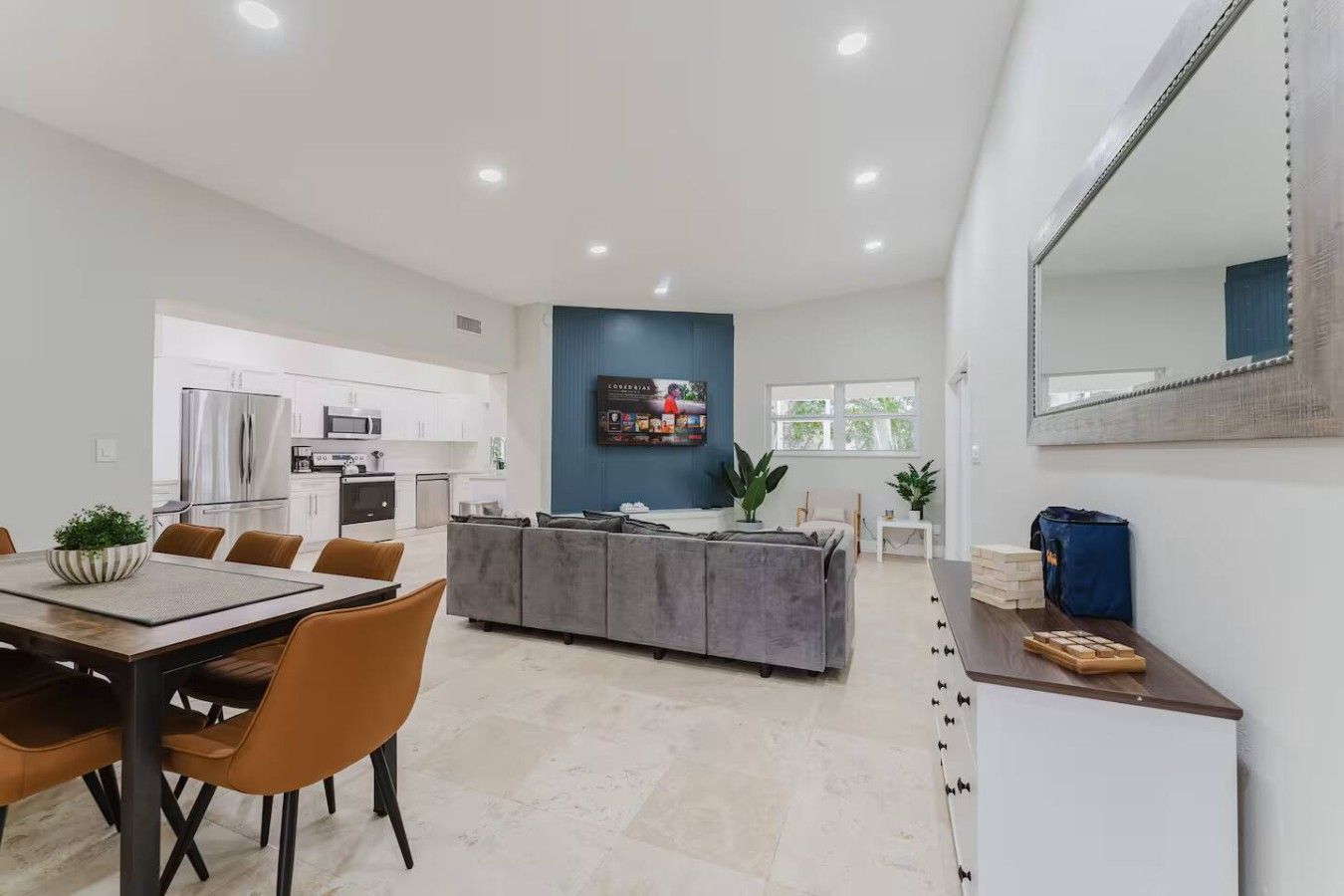 Una cocina moderna con electrodomésticos de acero inoxidable, un comedor con mesa y sillas de madera, y una sala de estar con sofá gris y un espejo grande.