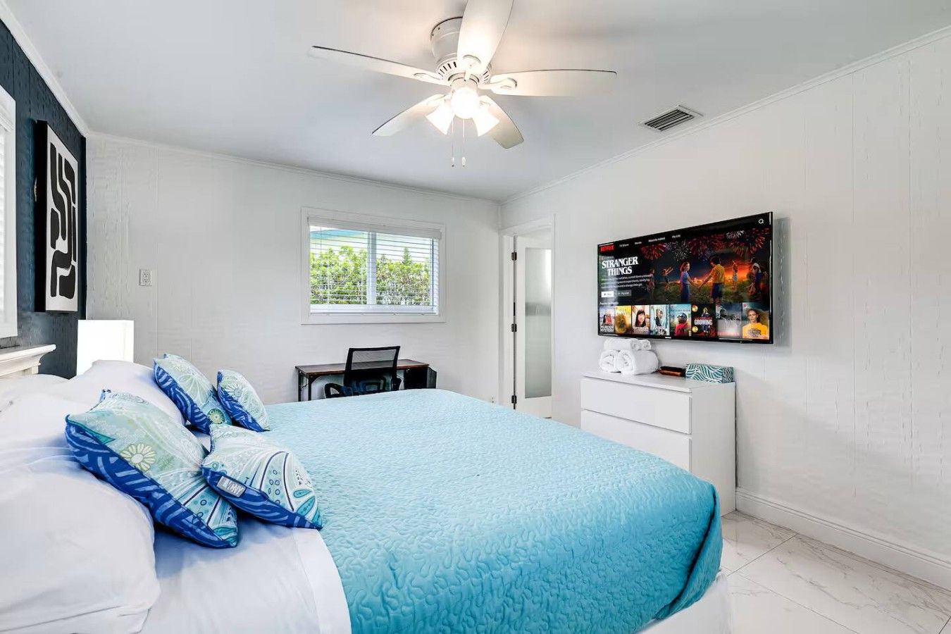 Ein Schlafzimmer mit Bett, Schreibtisch, Stuhl, Fernseher und Deckenventilator.