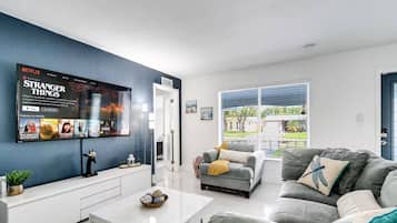 A modern living room with a flat-screen TV mounted on the wall, a grey sofa, a white coffee table, and a view of the outdoors through large windows.