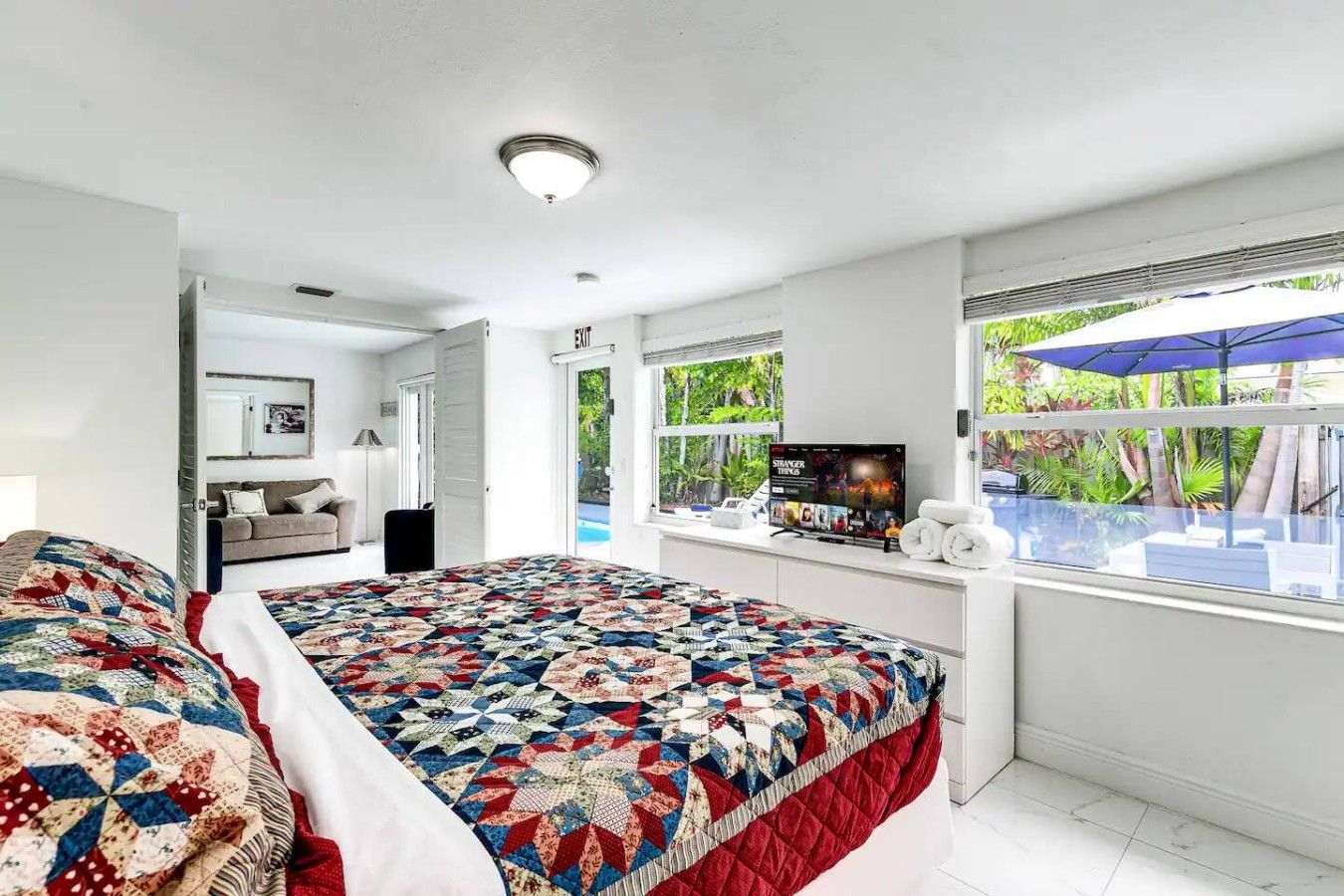 A hotel room with a large bed, a television, a window with a view of greenery, and a ceiling light.