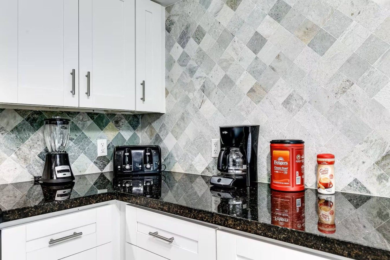 A kitchen with white cabinets, a granite countertop, a blender, a coffee maker, and two coffee containers.