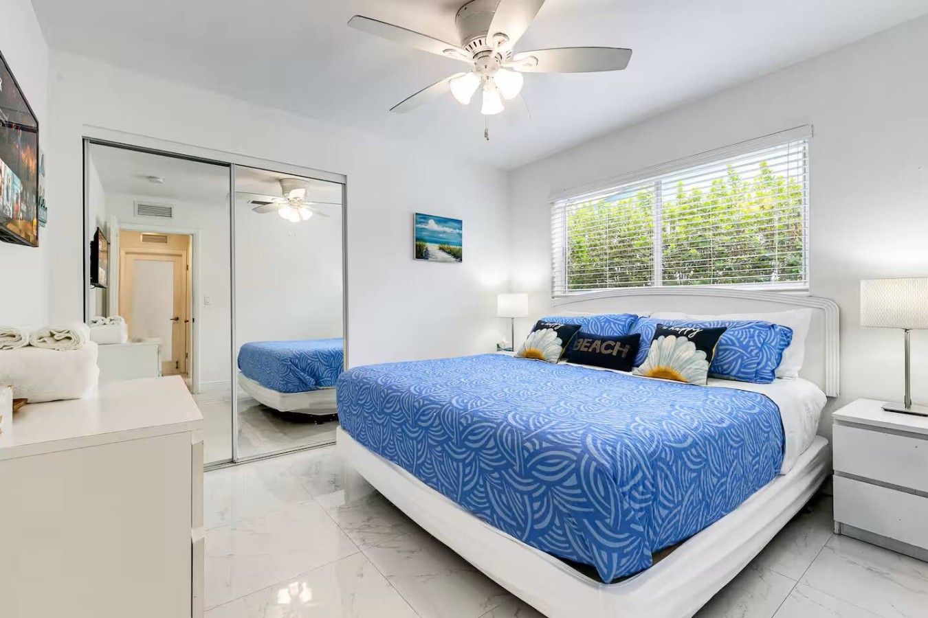 Quarto de hotel com cama grande, ventilador de teto, janela com persianas e espelho.
