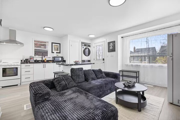 Una cocina moderna con armarios blancos, un comedor con sofá y una pequeña mesa, y un ventanal con vistas al exterior.