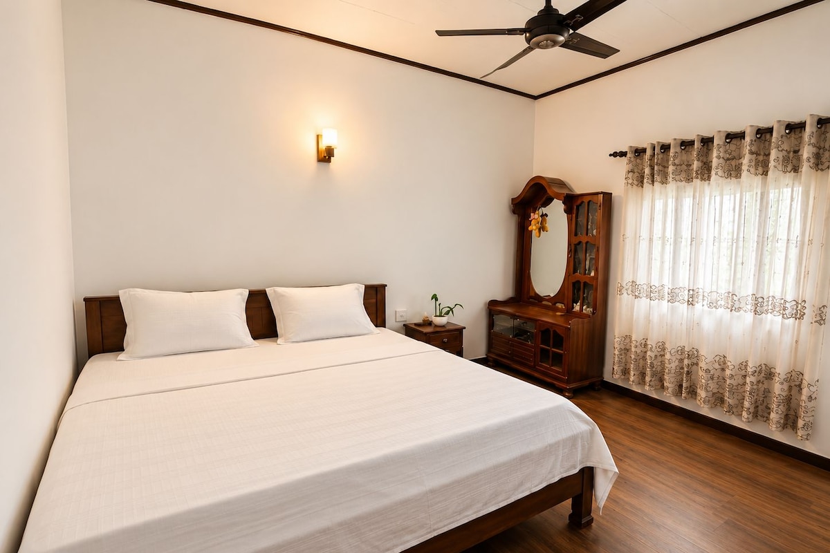 Quarto com cama, mesa de cabeceira, cômoda, ventilador de teto e janela com cortinas.
