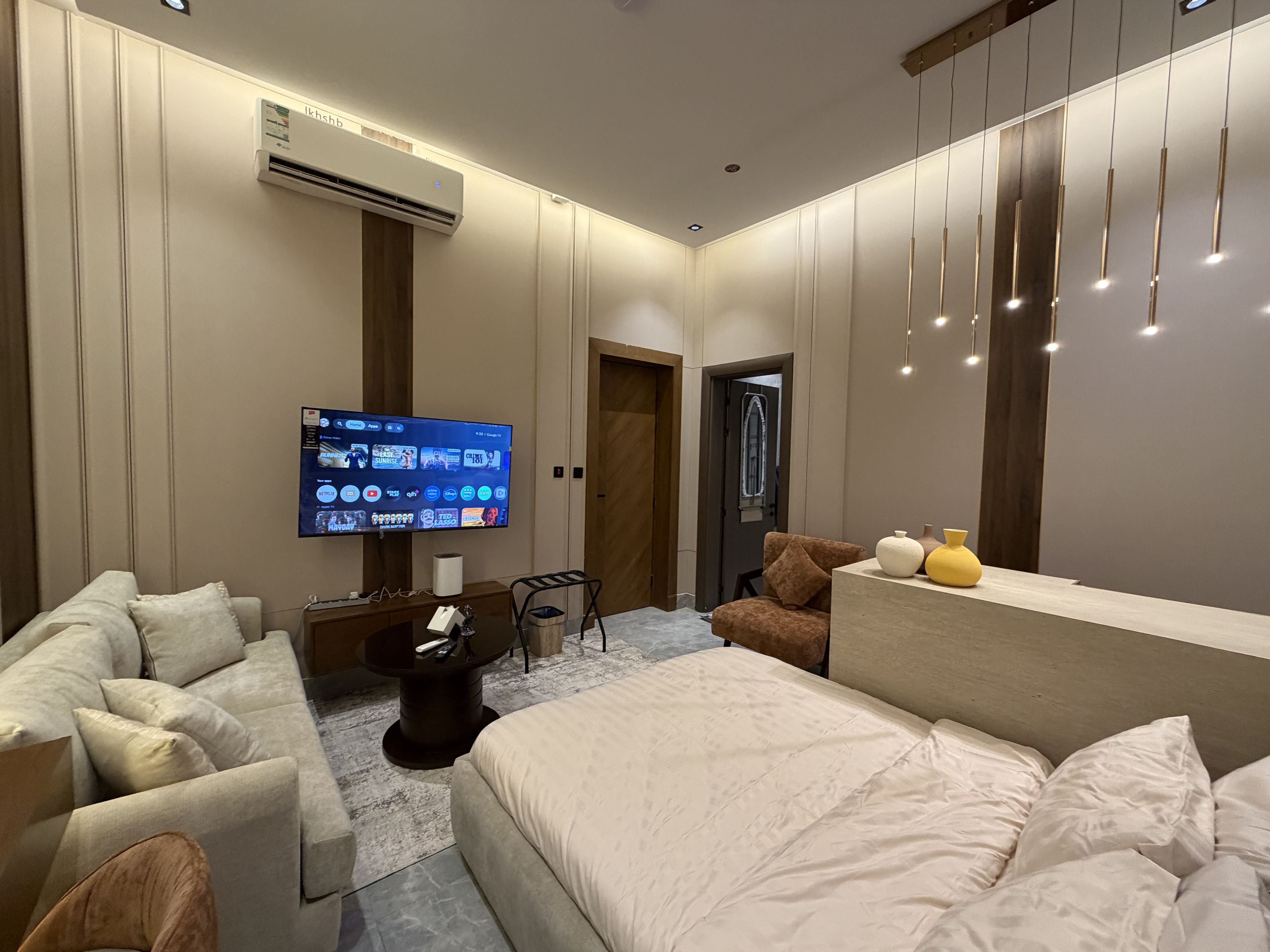 Quarto de hotel moderno com uma televisão de tela grande, um sofá, uma cama e uma pequena mesa com um cesto de lixo.
