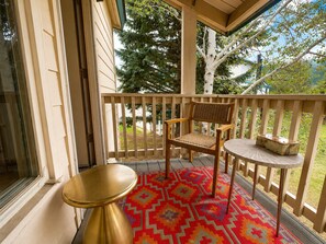Eine Holzterrasse mit einem Stuhl, einem kleinen Tisch, einem bunten Teppich und Blick auf Bäume sowie ein Haus im Hintergrund.