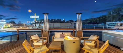 En terrasse med udendørs møbler og bålsted, med udsigt over en lysthavn med både og et skilt til en restaurant.