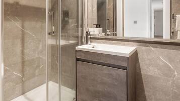 Een moderne badkamer met een douchecabine van glas, een wit wastafeltje en een wand betegeld met marmer.