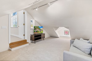 A modern attic bedroom with a sloped ceiling, a wooden entertainment unit, a gray sofa, and a large window with a view of greenery.