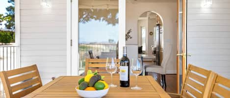 Una mesa de madera con un cuenco de fruta y una botella de vino, frente a una pared blanca con una puerta que da a un balcón.