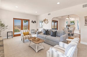 A spacious living room with a large grey sofa, striped armchairs, and a coffee table. There is a sliding glass door leading to a balcony with a view of the countryside.