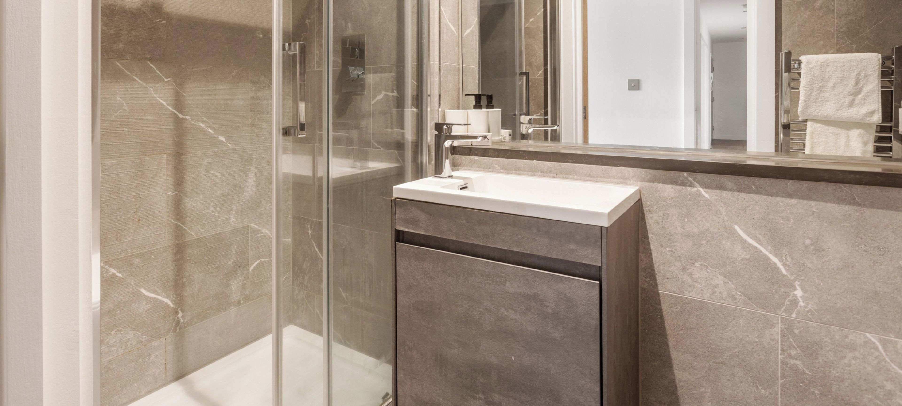 Een moderne badkamer met een douchecabine van glas, een witte wastafel en een wand betegeld met marmer.