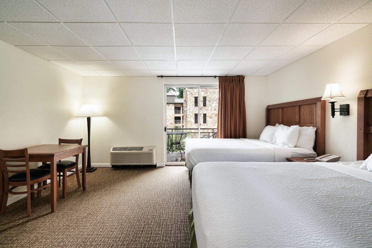 Habitación de hotel con dos camas, un escritorio con silla, una lámpara y vistas a un edificio a través de la ventana.